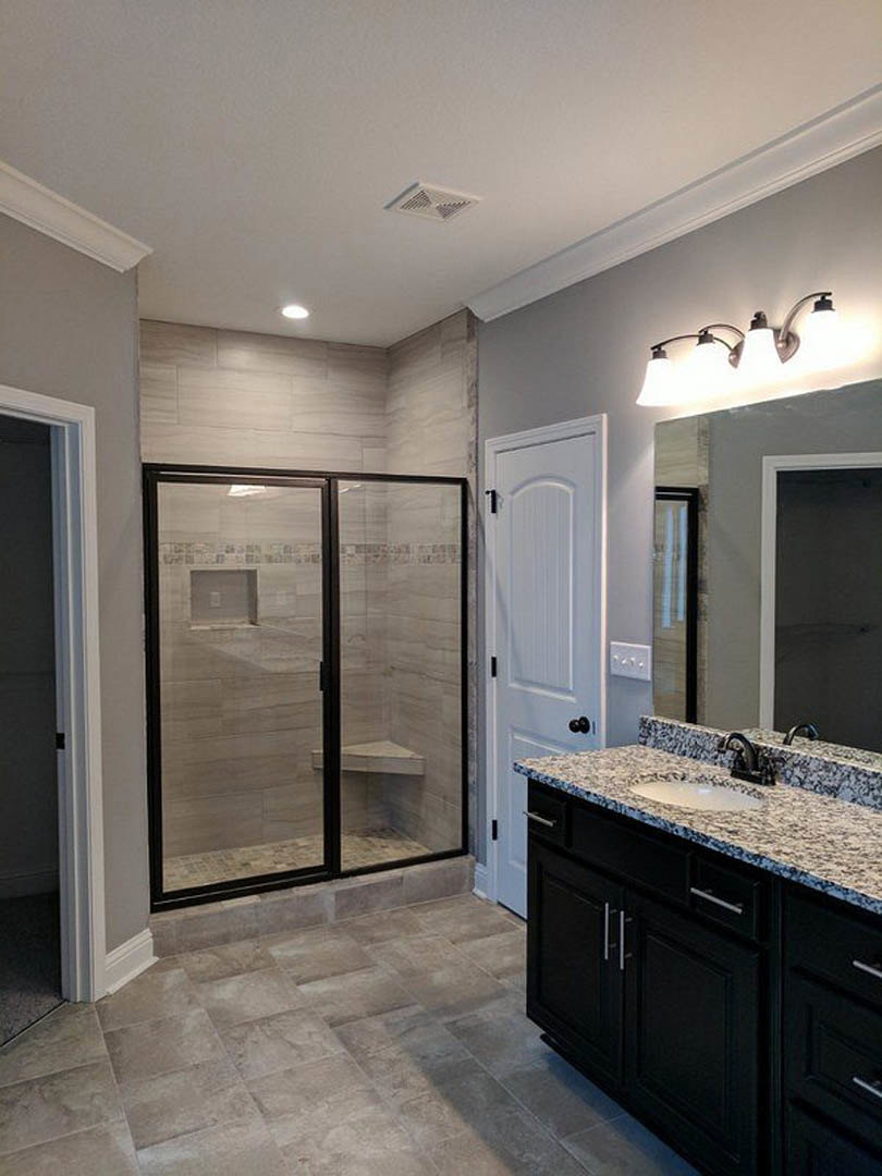 Bathroom with glass-enclosed shower, black vanity topped with marble, white sink, wall-mounted row of lights, white door, and tile flooring.