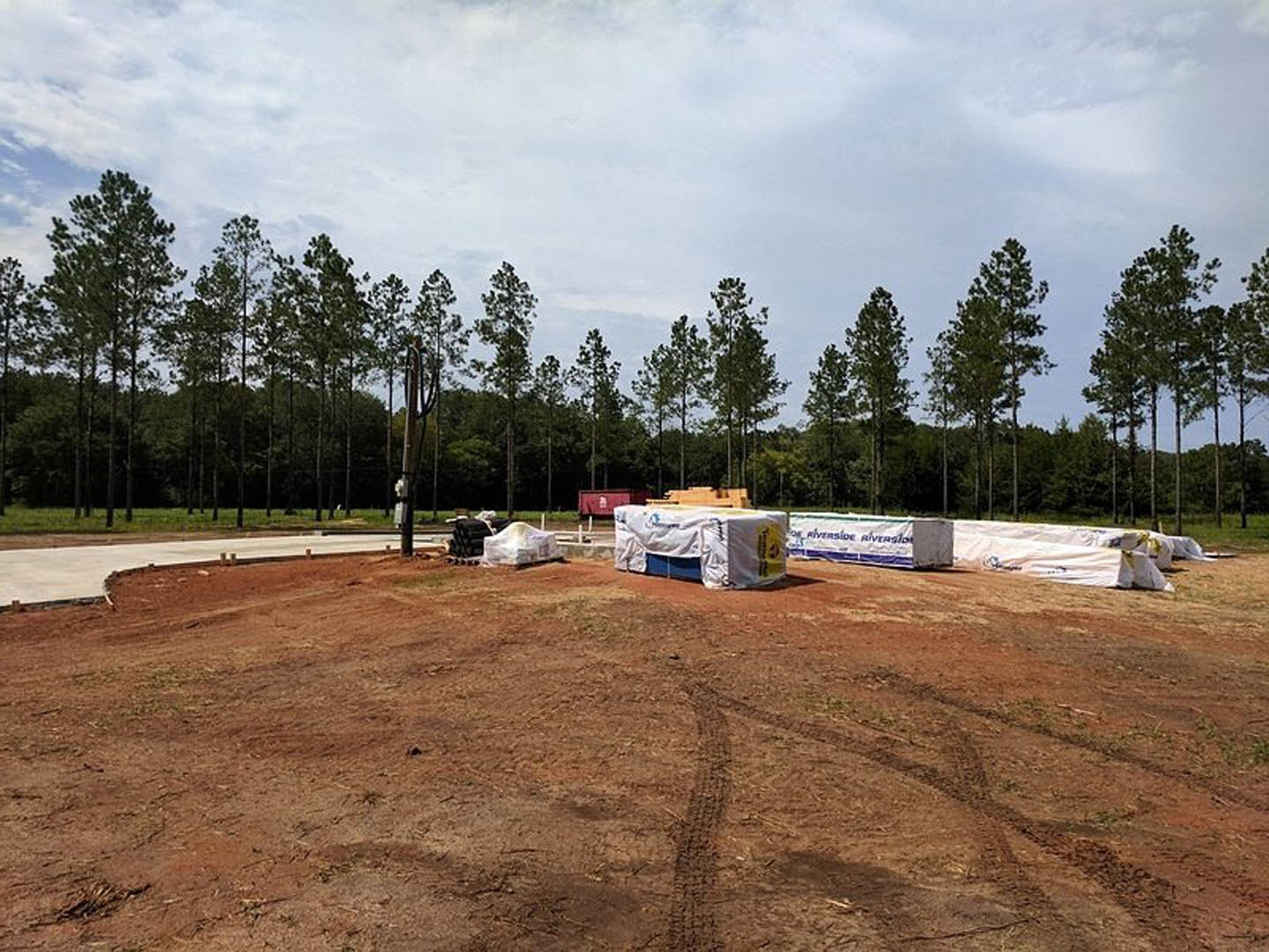 Expansive dirt lot with tire tracks, bordered by a group of trees and a distant building, under a partly cloudy sky