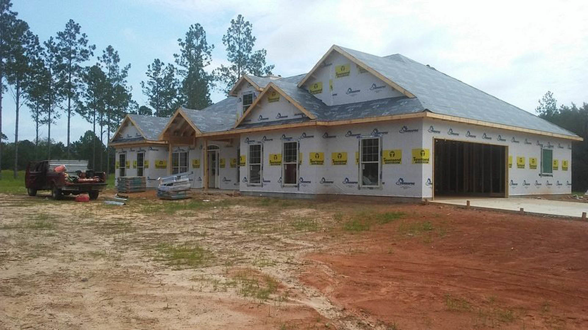 Two-story house under construction with exposed framing, yellow and white signage, dirt-covered yard, scattered grass, and a wooden fence; construction truck visible in background
