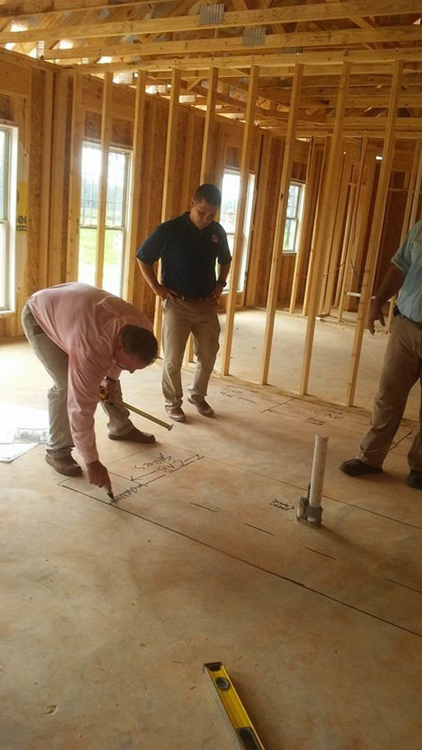 Two men measuring and discussing a floor plan drawn on unfinished wood flooring, one holding a tape measure and the other with hands in pockets, surrounded by construction tools