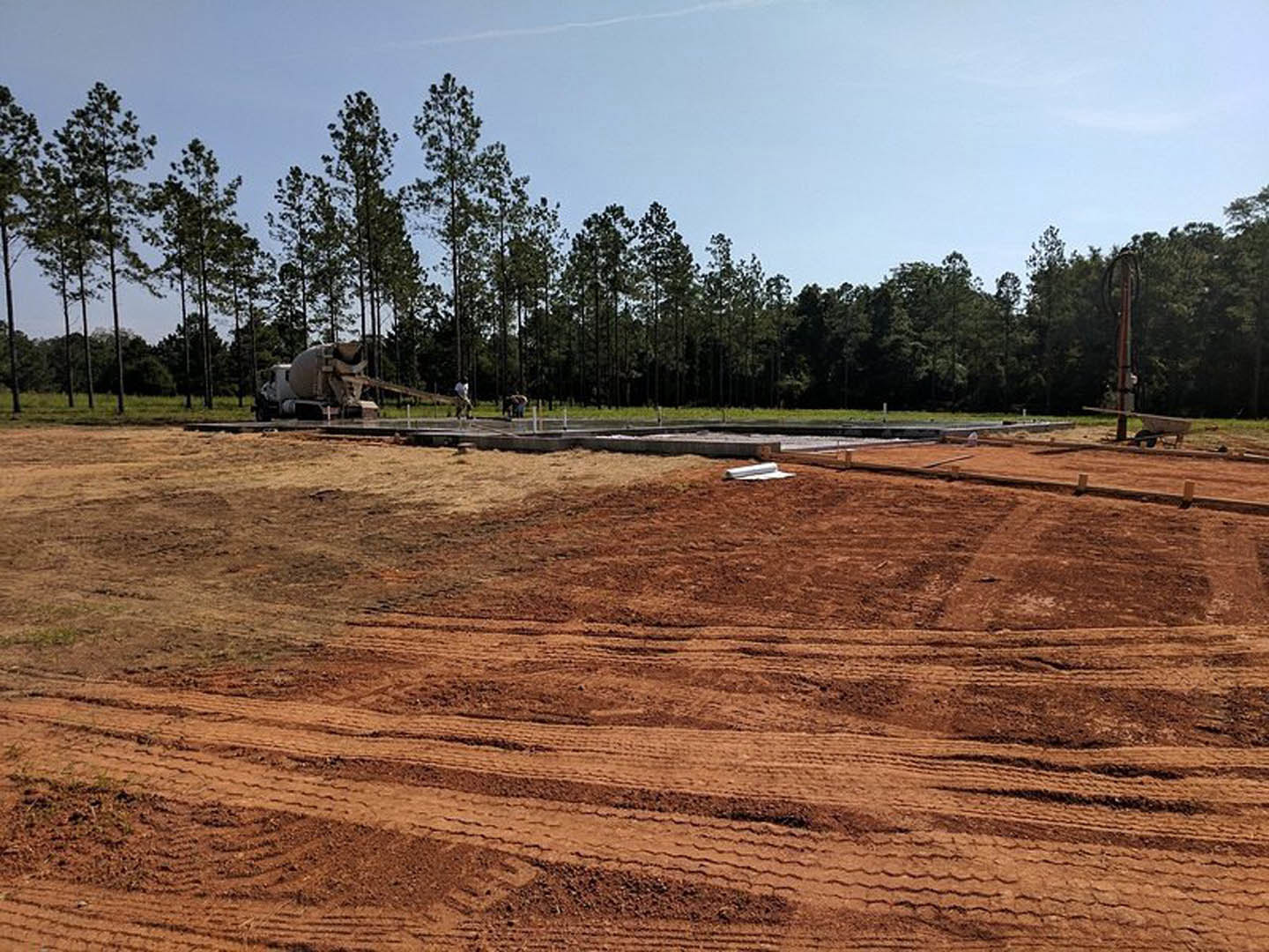 Dirt construction site with tire tracks, concrete mixer truck, grassy land lot, and dense trees in the background under a partly cloudy sky