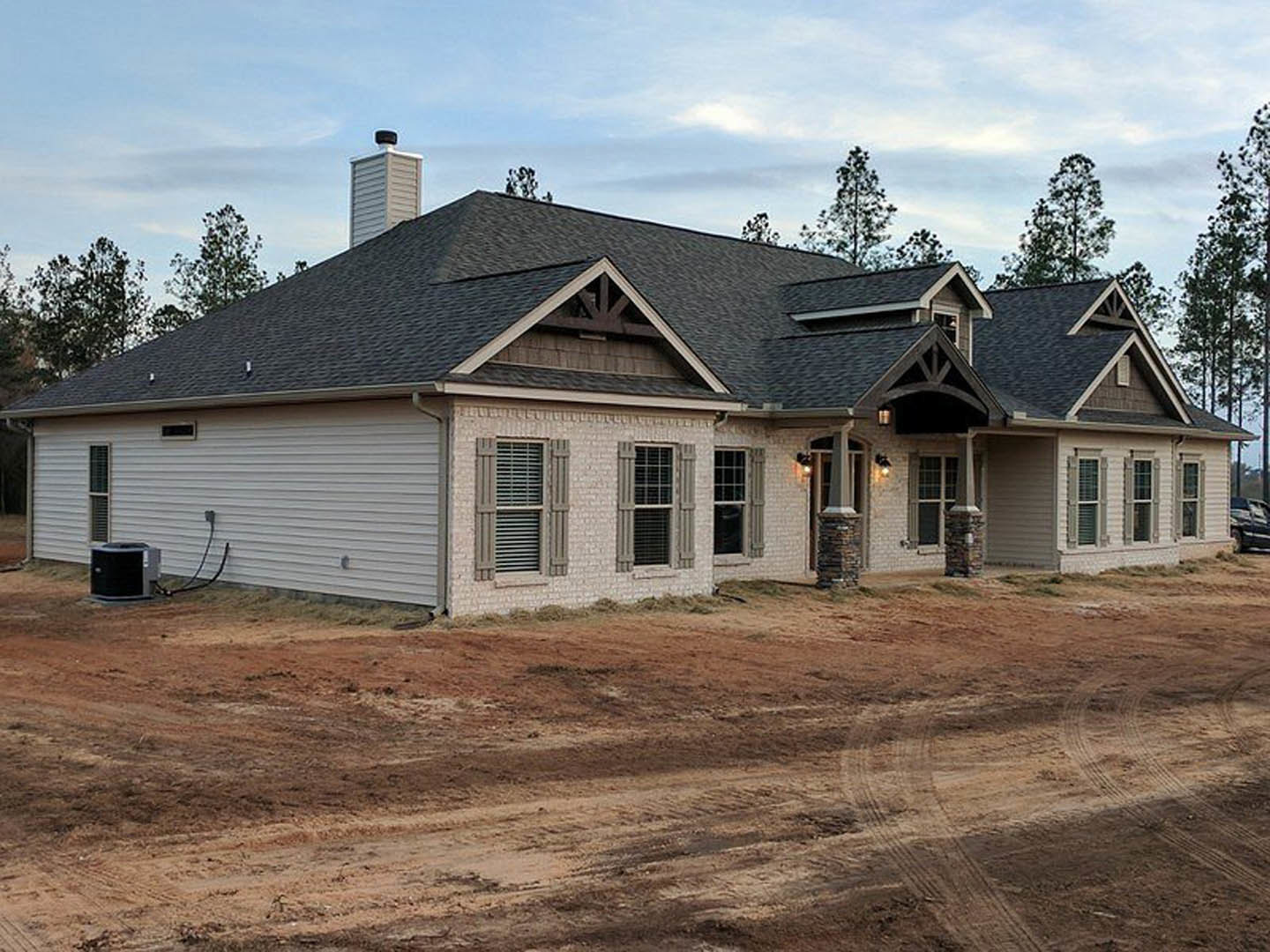 Single-story house with light-colored siding, dark shingle roof, barred window, dirt driveway, and sparse landscaping; black utility box on ground near entrance.