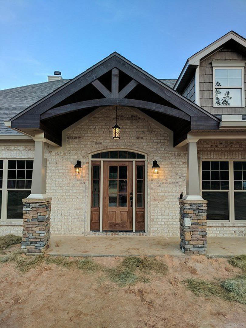 Wood front door framed by stone column, large window reflecting tree, light-colored siding, and landscaped entry.
