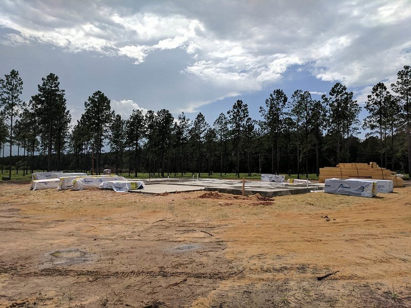 Partially built custom home framed with exposed wood, construction materials scattered on dirt lot, tall trees lining background, cloudy sky overhead
