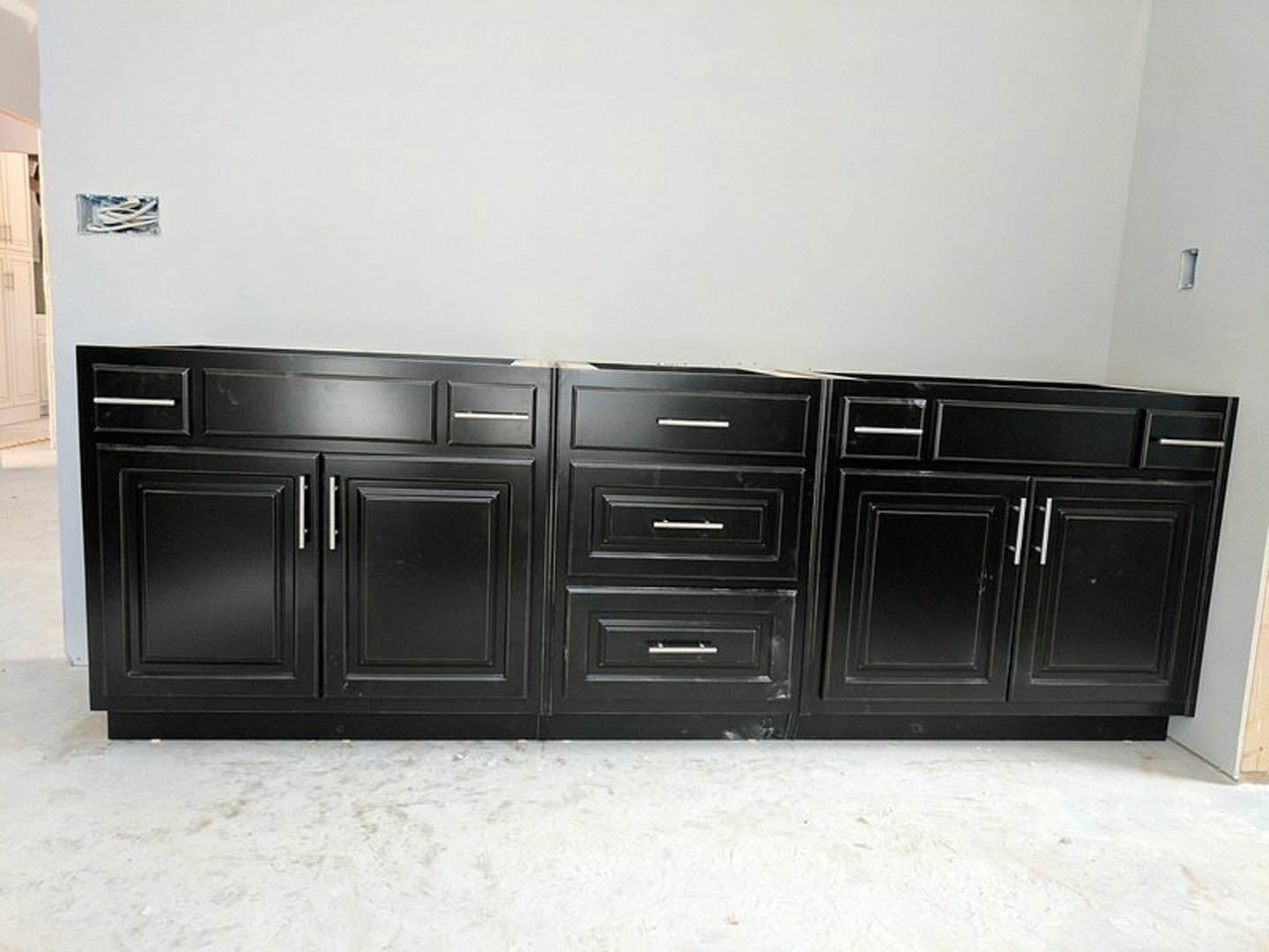 Black kitchen cabinets with silver handles against a white wall, light-colored flooring, and modern cabinetry.