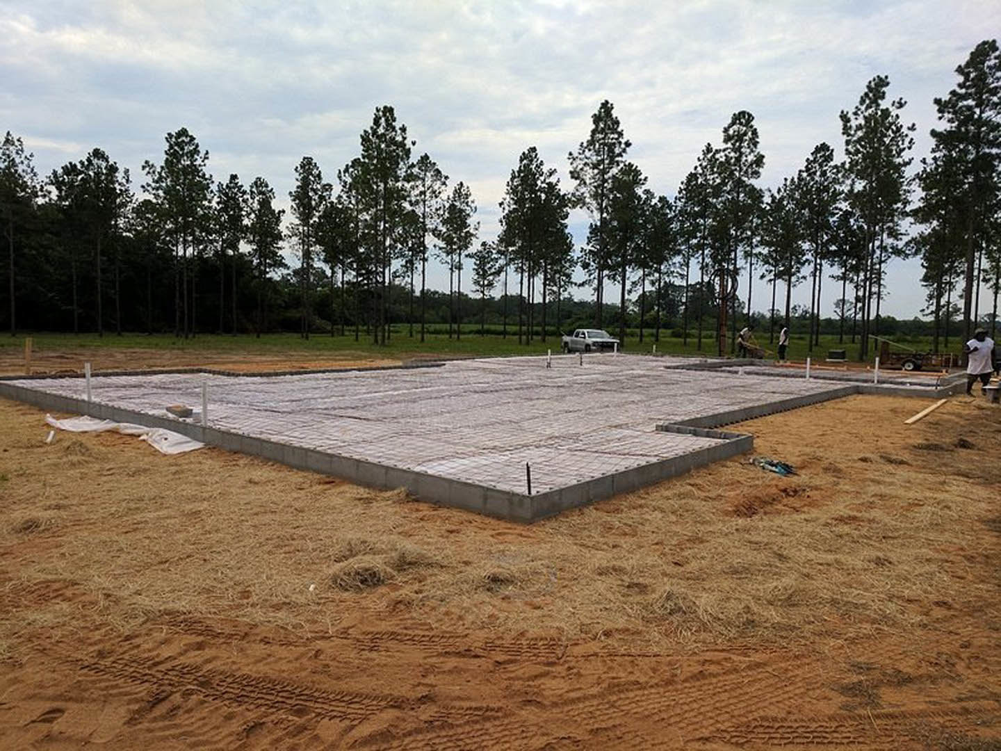 Concrete foundation slab set in a grassy field with trees and a parked car in the background; person in white shirt near the edge; white plastic bag on dirt close to the slab.