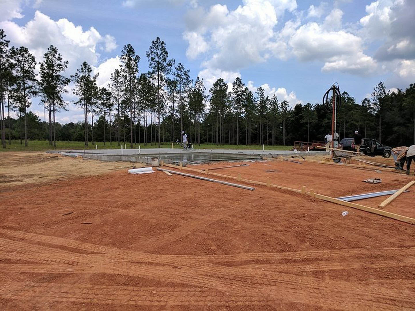 Construction site with dirt ground, tire tracks, scattered grass, black truck parked nearby, red and white pole, and surrounding trees under a blue sky.