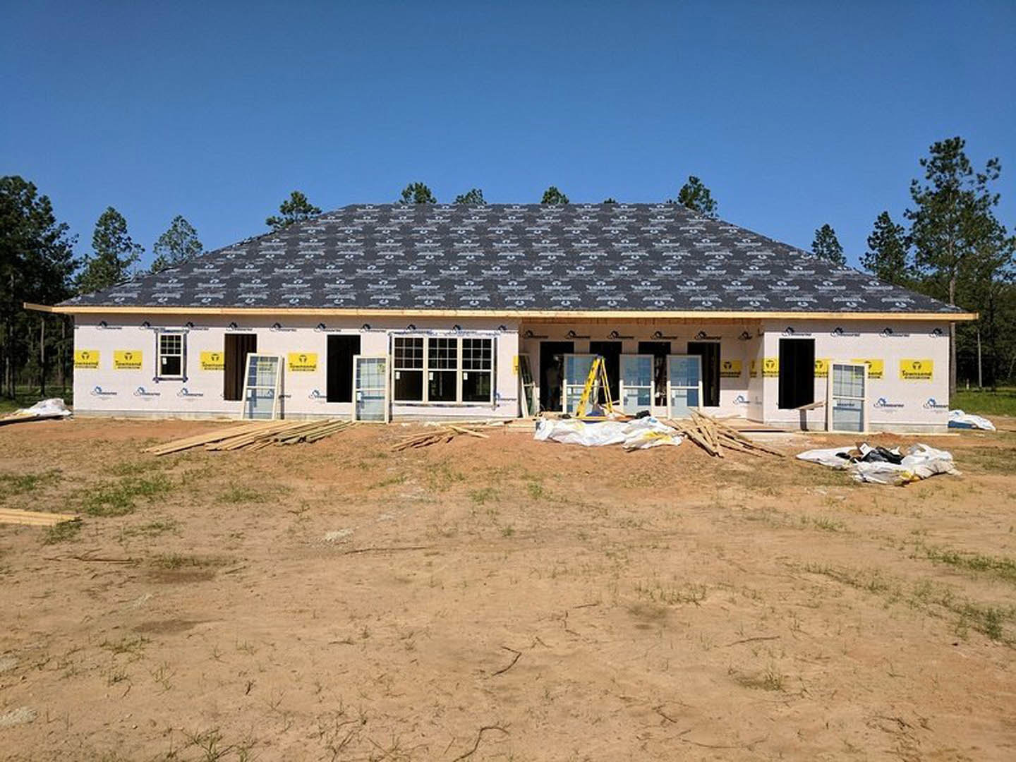 Framed wooden house under construction with roof installed, exposed beams and plywood, dirt yard in foreground, black door with white trim, window close-up, trees and sky in
