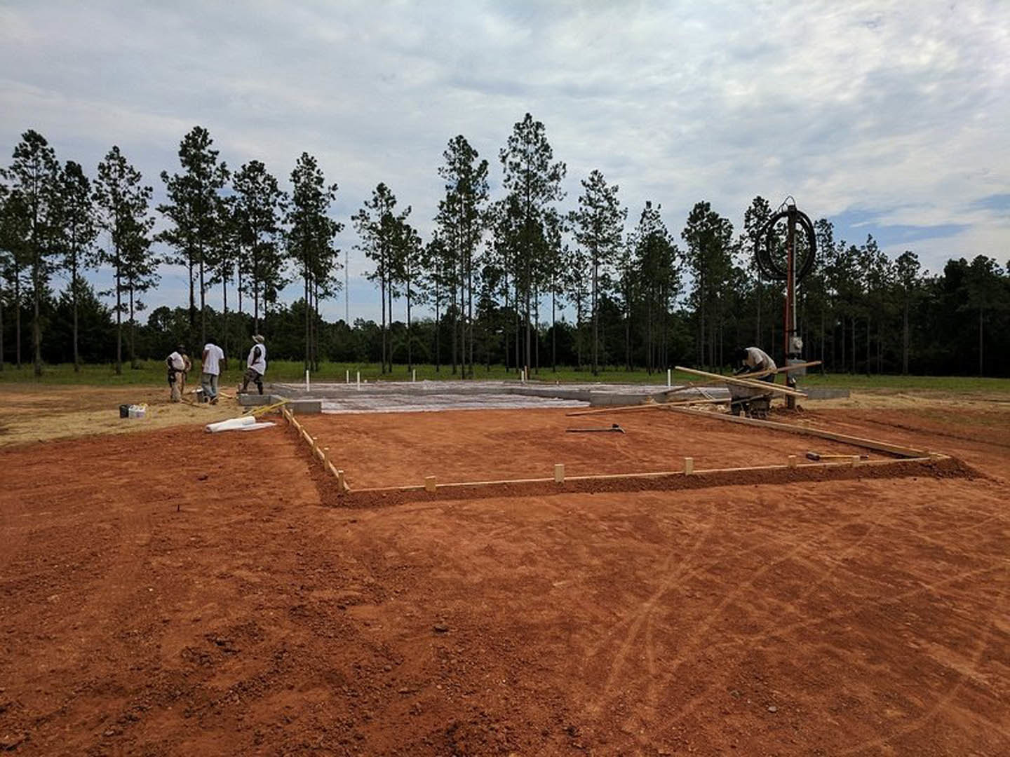 Framed wood structure under construction with workers assembling beams, surrounded by dirt and grass, trees in the background, cloudy sky overhead