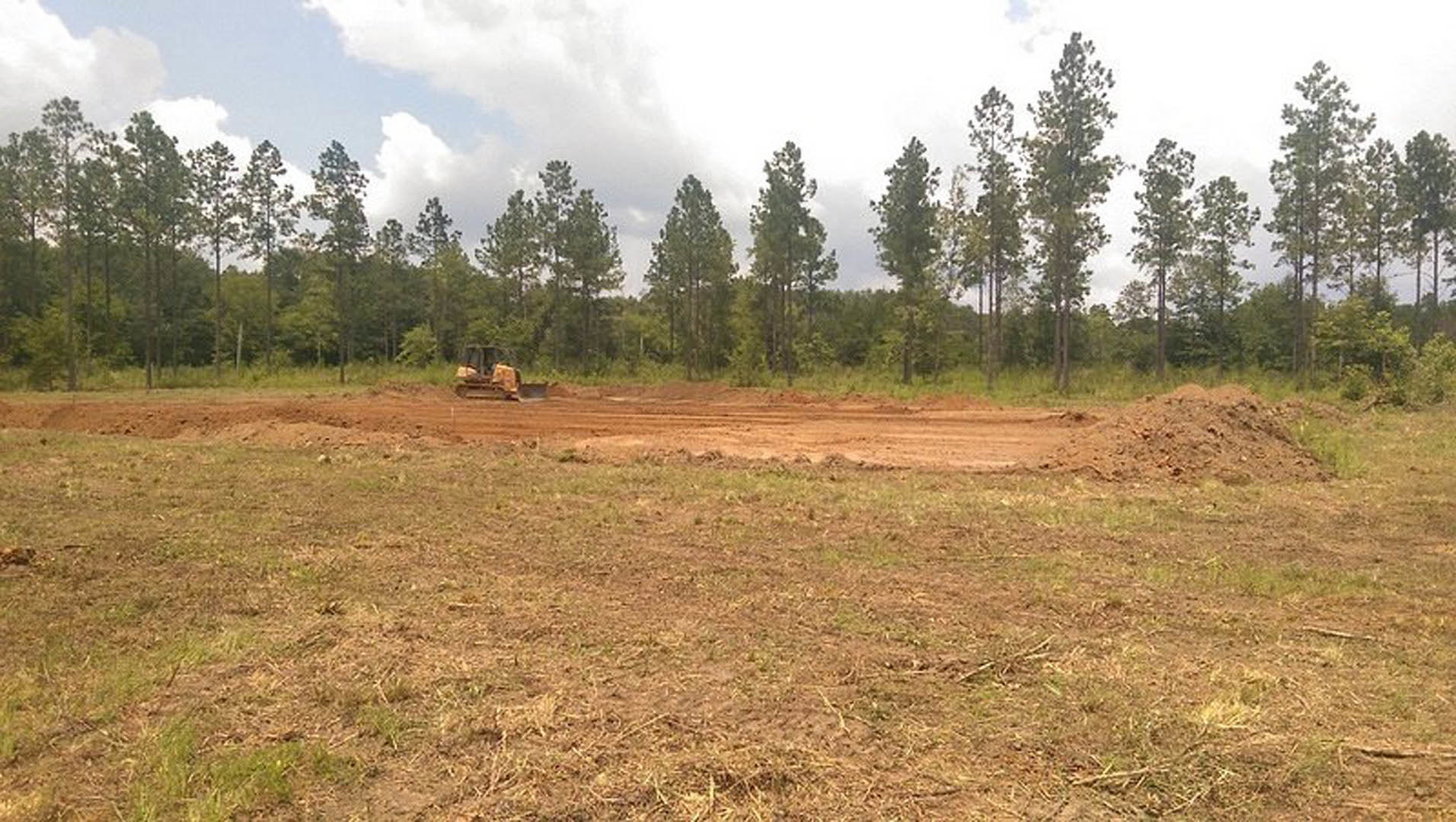 Yellow bulldozer clearing grassy dirt field with scattered trees under cloudy sky