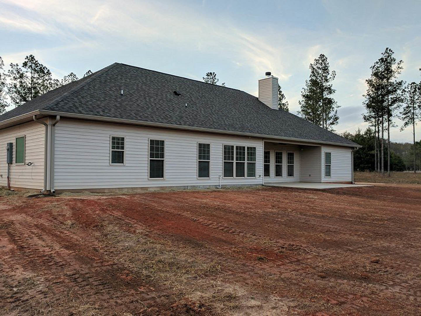 Tall white house with black roof, white-framed windows, and dirt yard in foreground; Wyckoff House and trees visible in background under cloudy sky.