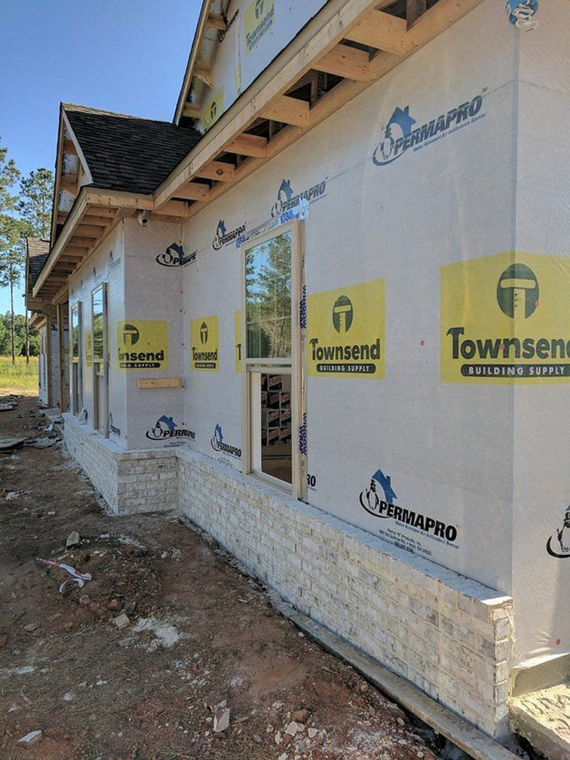 Partially built brick exterior with glass window and door, yellow construction sign with black text, unfinished roof, and company logo displayed on side