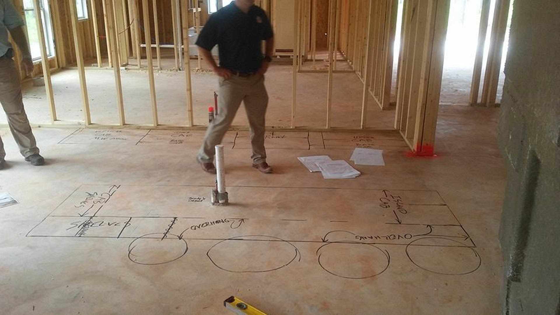 Man in black shirt and jeans standing on unfinished wood floor in residential construction site, surrounded by exposed framing and construction materials.