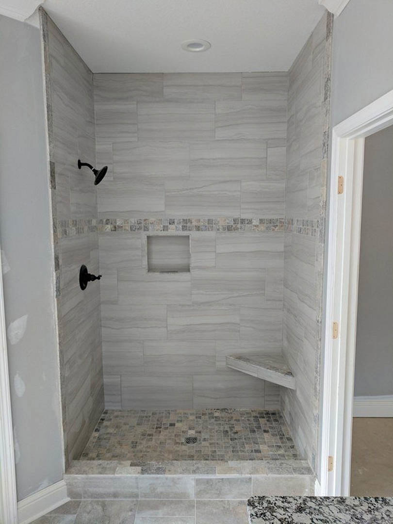 Walk-in shower with light gray tile floor, round metal drain, white plaster walls, chrome shower head, and black soap dish on built-in shelf
