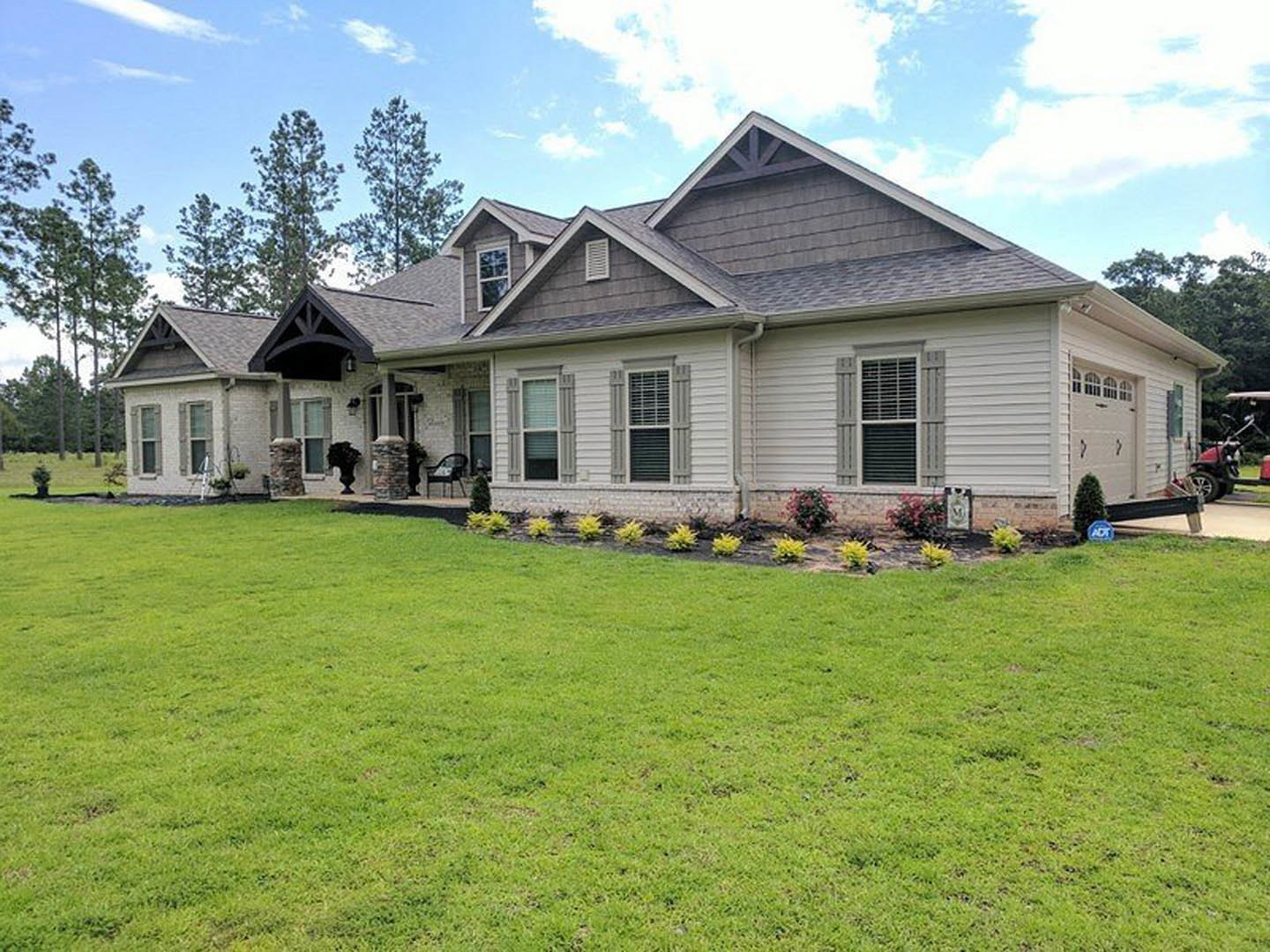 Two-story farmhouse with white siding, black shutters, and front porch, surrounded by green lawn, bushes, and mature trees; a car parked in driveway and outdoor chair near