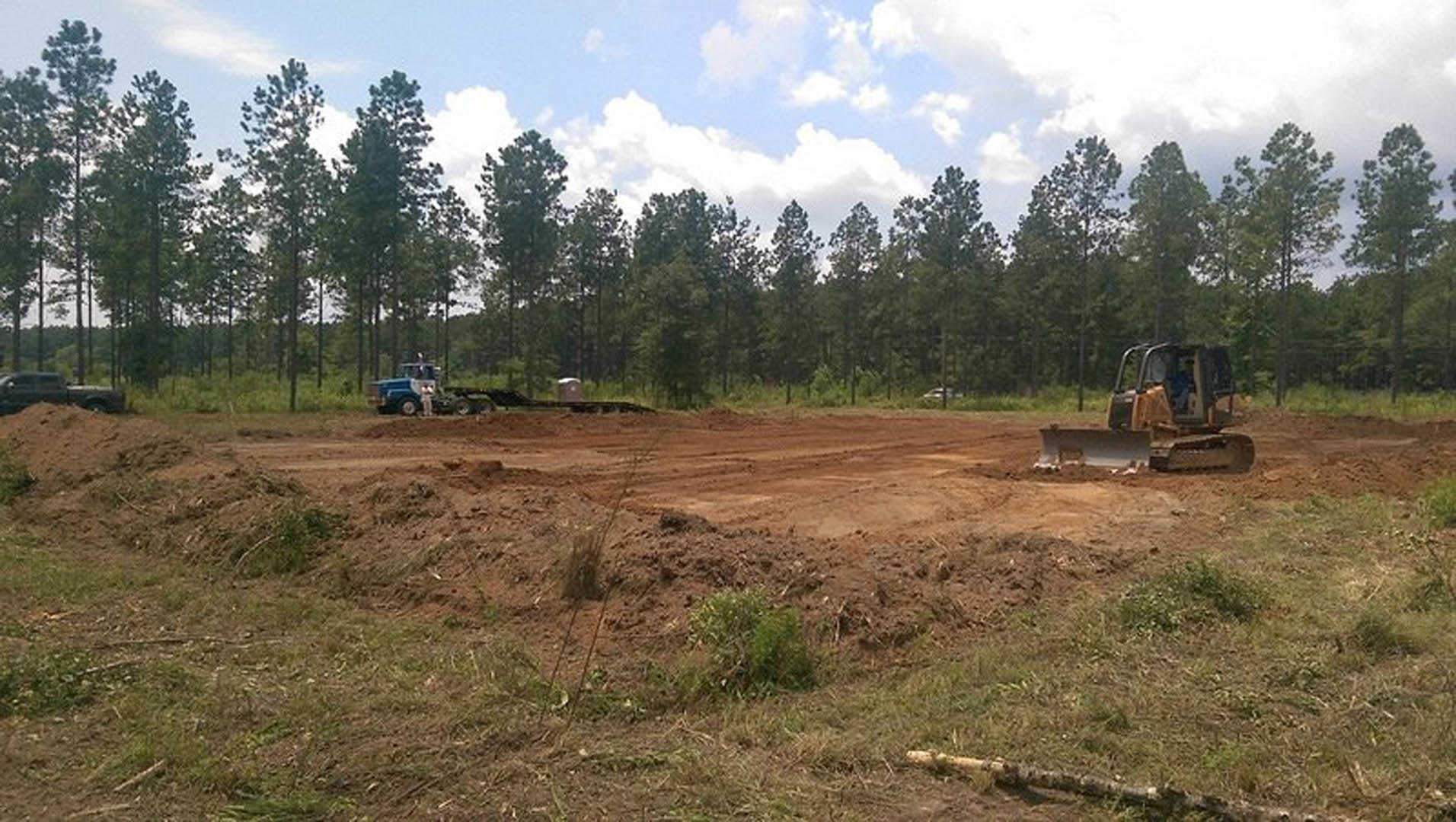 Tractor and bulldozer parked on a dirt field with a fence, surrounded by grassy land and trees under a partly cloudy blue sky