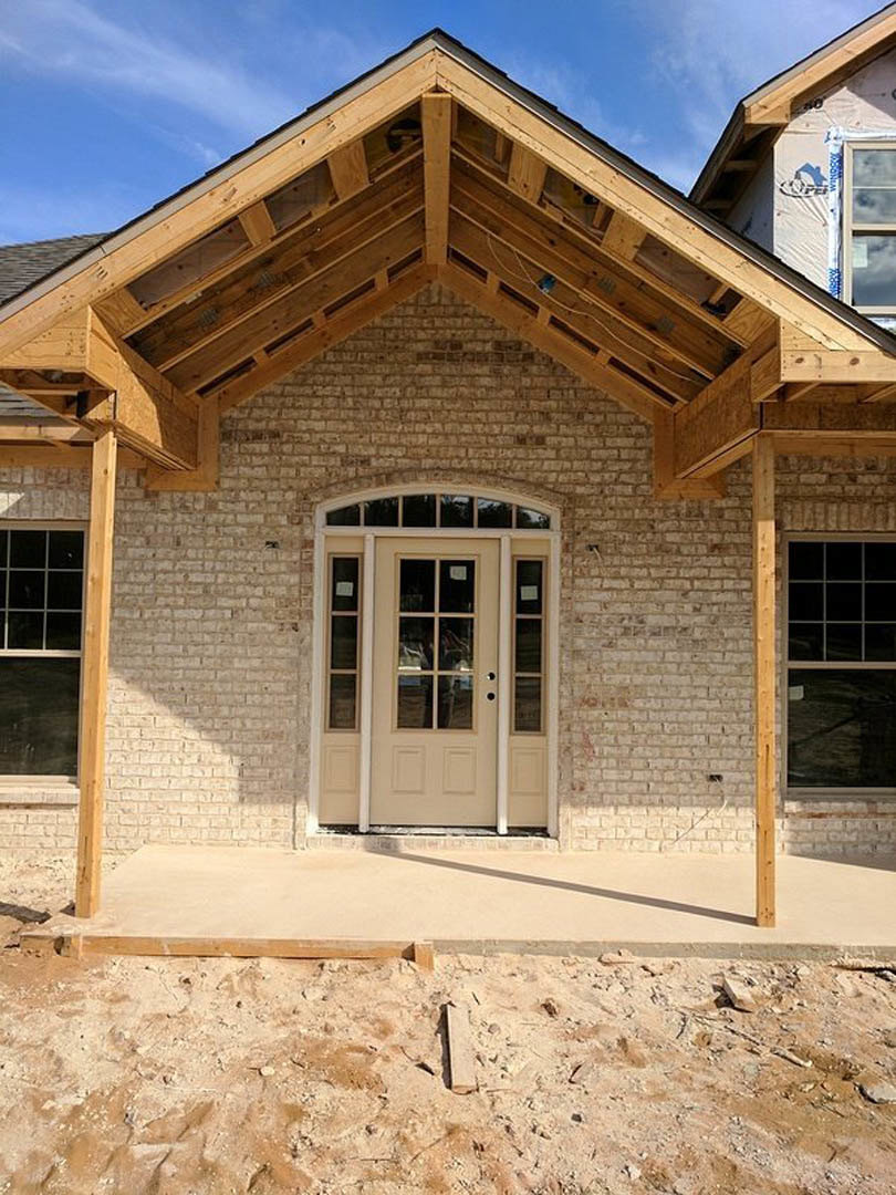 Partially built house with shingled roof, exposed wooden framing, square window, front porch, close-up door, and concrete surface showing shadow.