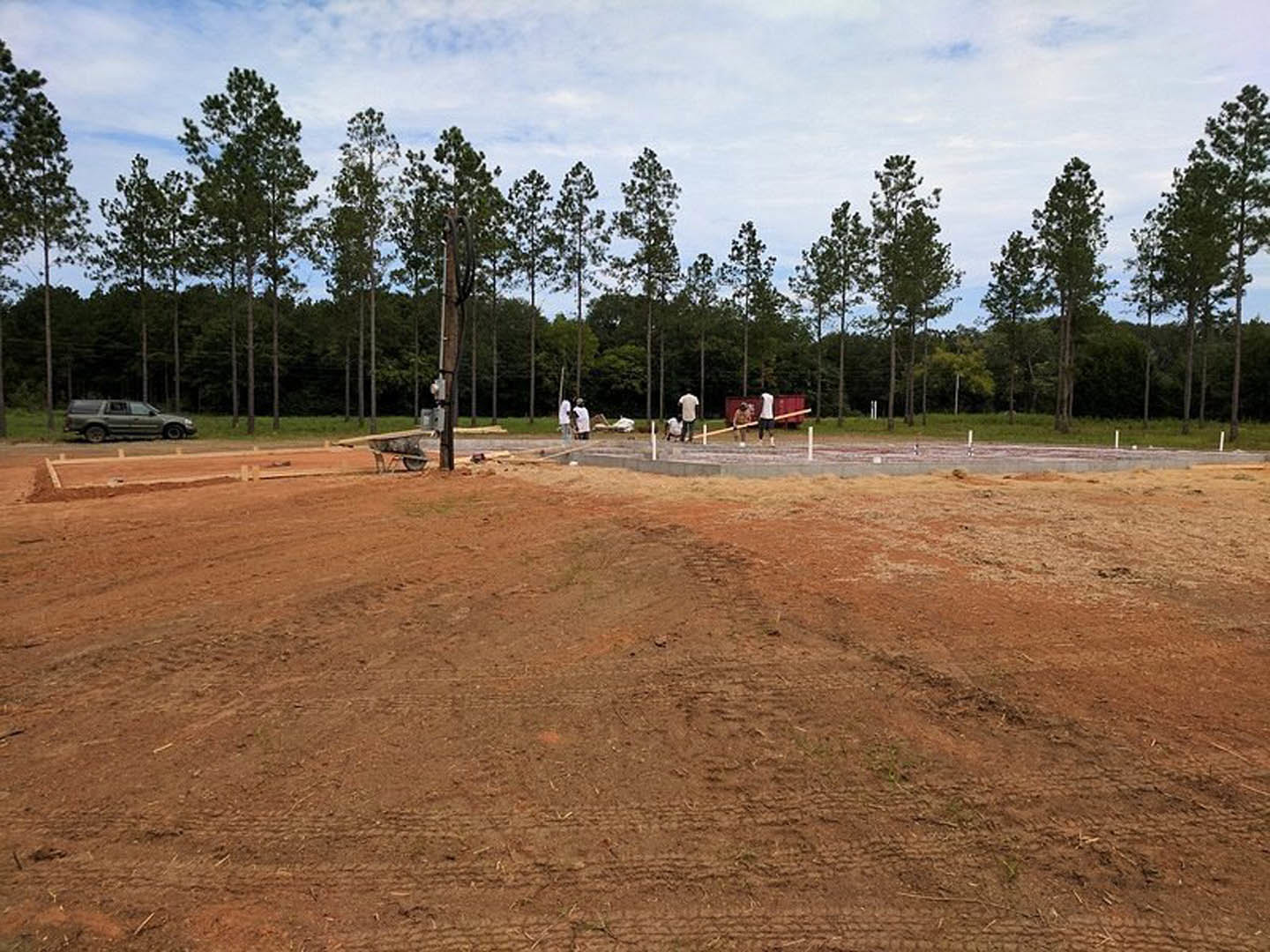 Dirt construction site bordered by tall trees, people standing near a wood utility pole, car parked on roadside, cloudy sky overhead