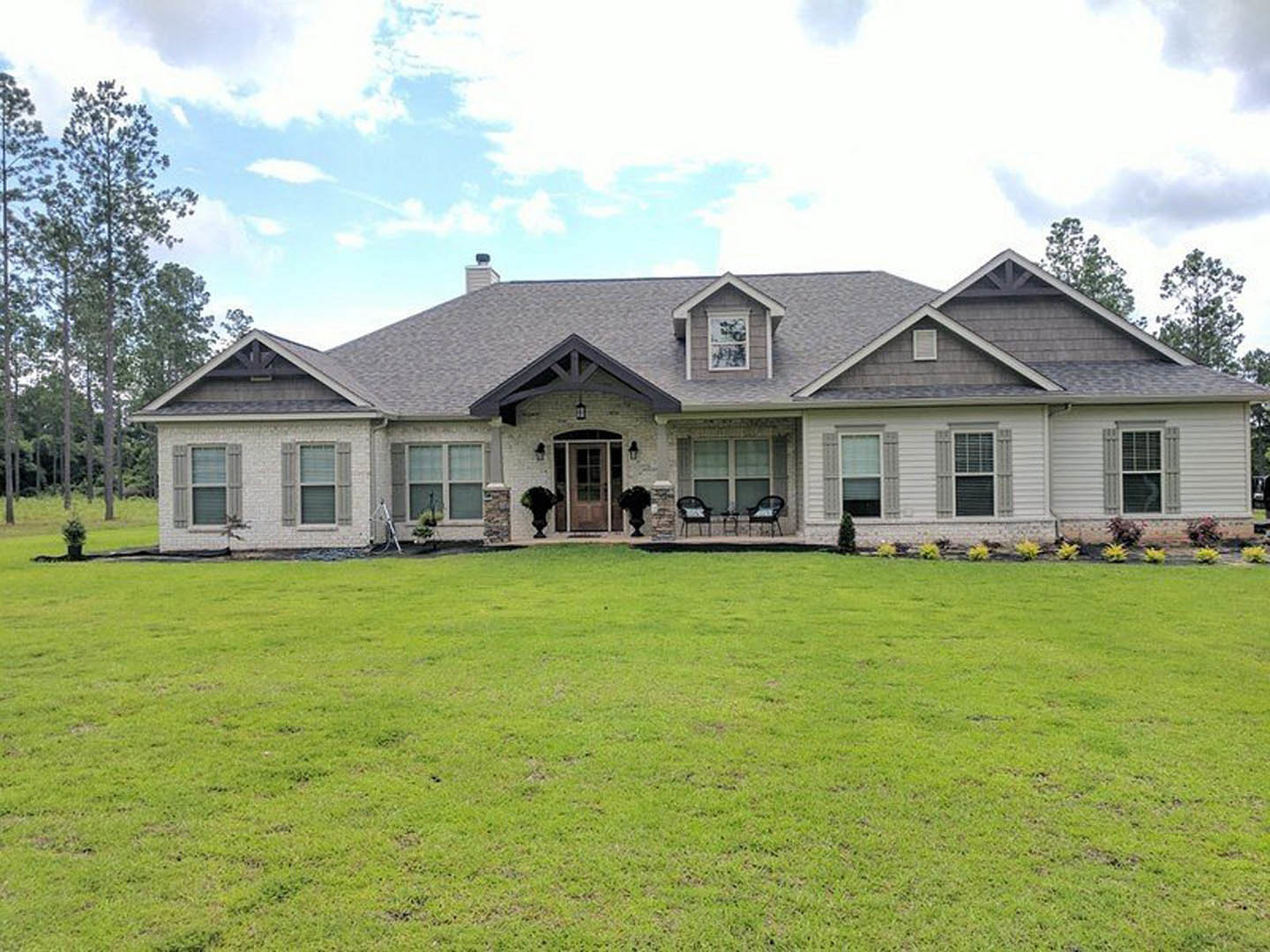 White farmhouse with covered porch, dark front door, large windows, manicured green lawn, outdoor chairs, and tall tree in foreground under partly cloudy sky