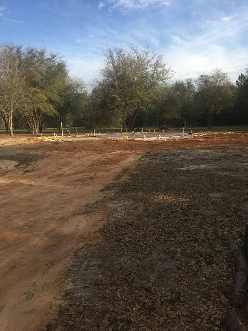 Dirt road bordered by a wooden fence, mature leafy trees under blue sky with scattered clouds