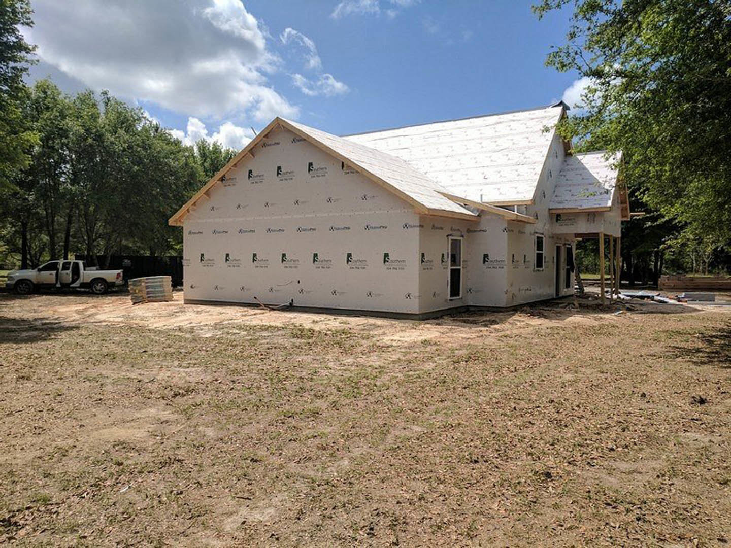 Wood-framed house under construction with exposed roof trusses, surrounded by tall trees, dirt lot in foreground, blue sky with scattered clouds overhead, white van parked nearby