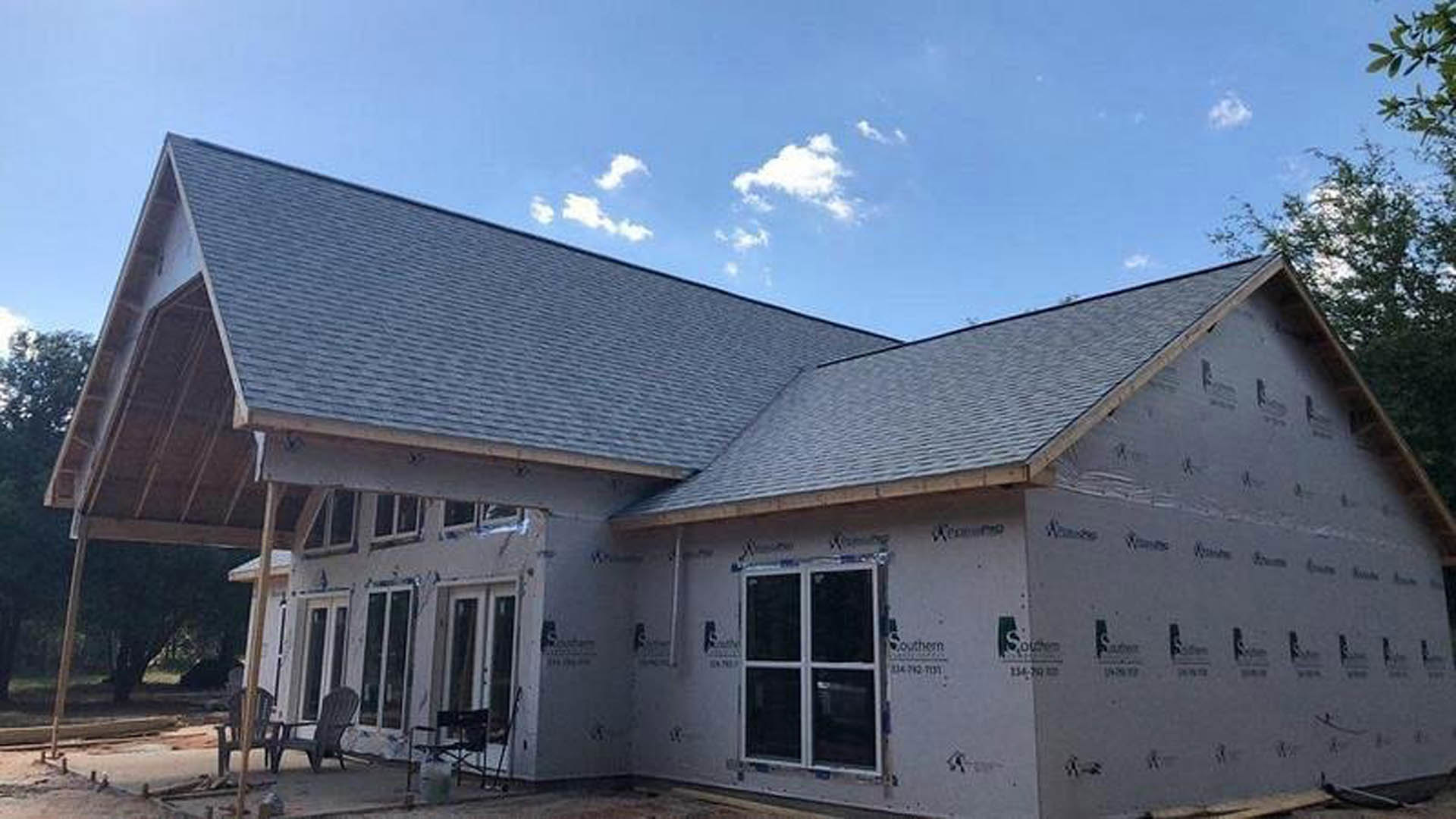 Framed house under construction with exposed wooden siding, white window frames, and unfinished roof beneath clear blue sky