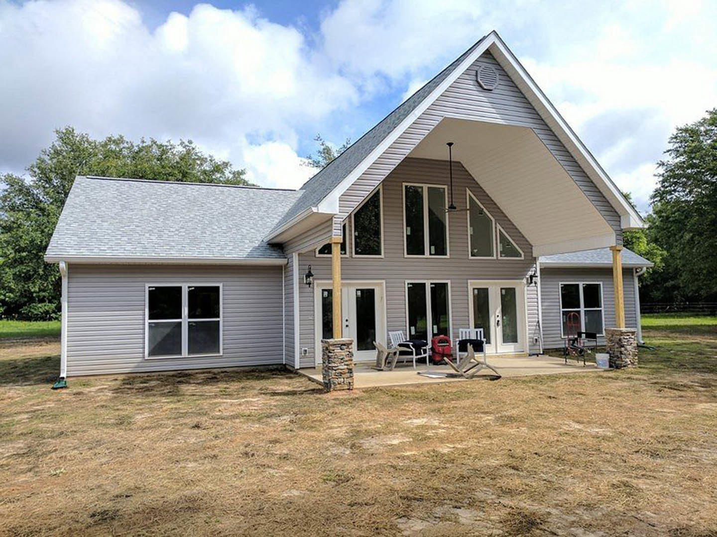 Modern two-story house with large windows, covered patio featuring outdoor seating, manicured green lawn, and mature trees under a partly cloudy sky