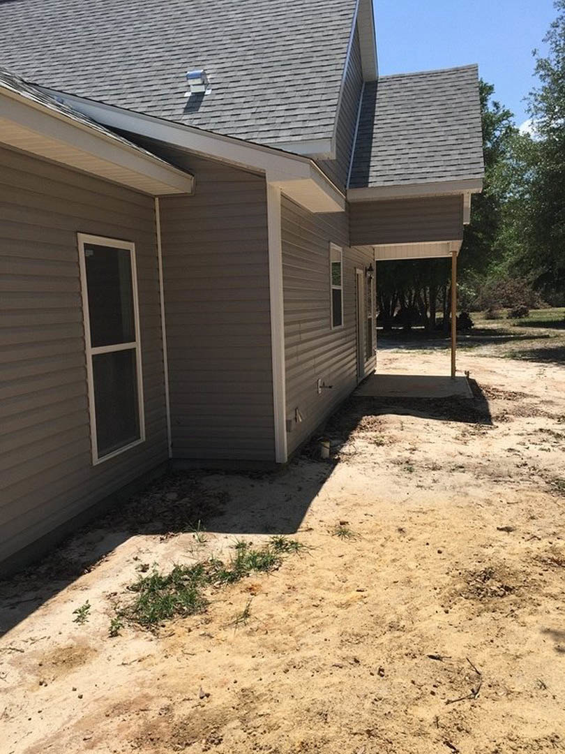 Two-story home with light siding, large windows, and a dirt yard bordered by a concrete driveway and sparse landscaping.