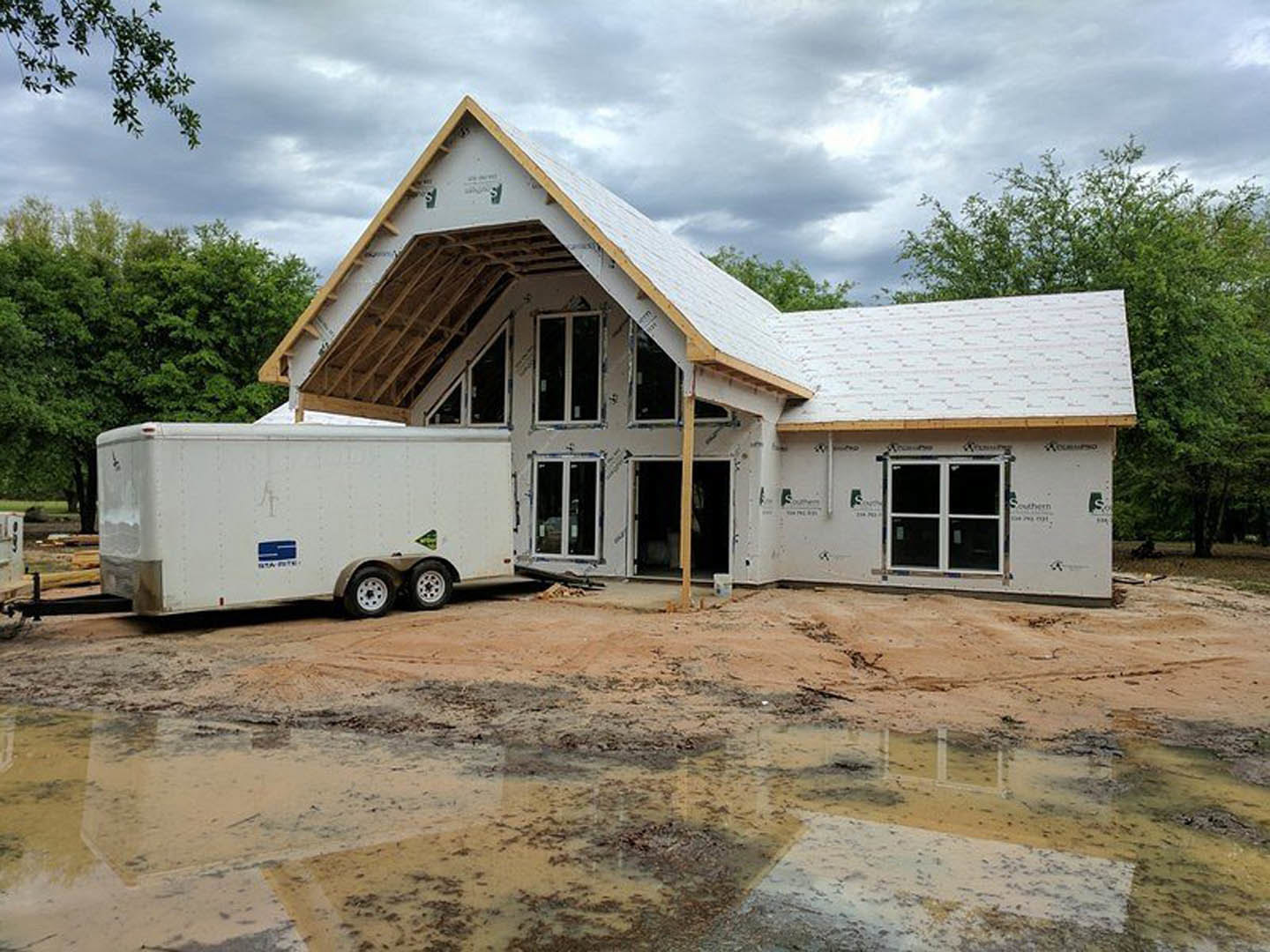 Framed custom home under construction with exposed wood, large windows, and a utility trailer parked on dirt lot; cloudy sky and trees in background