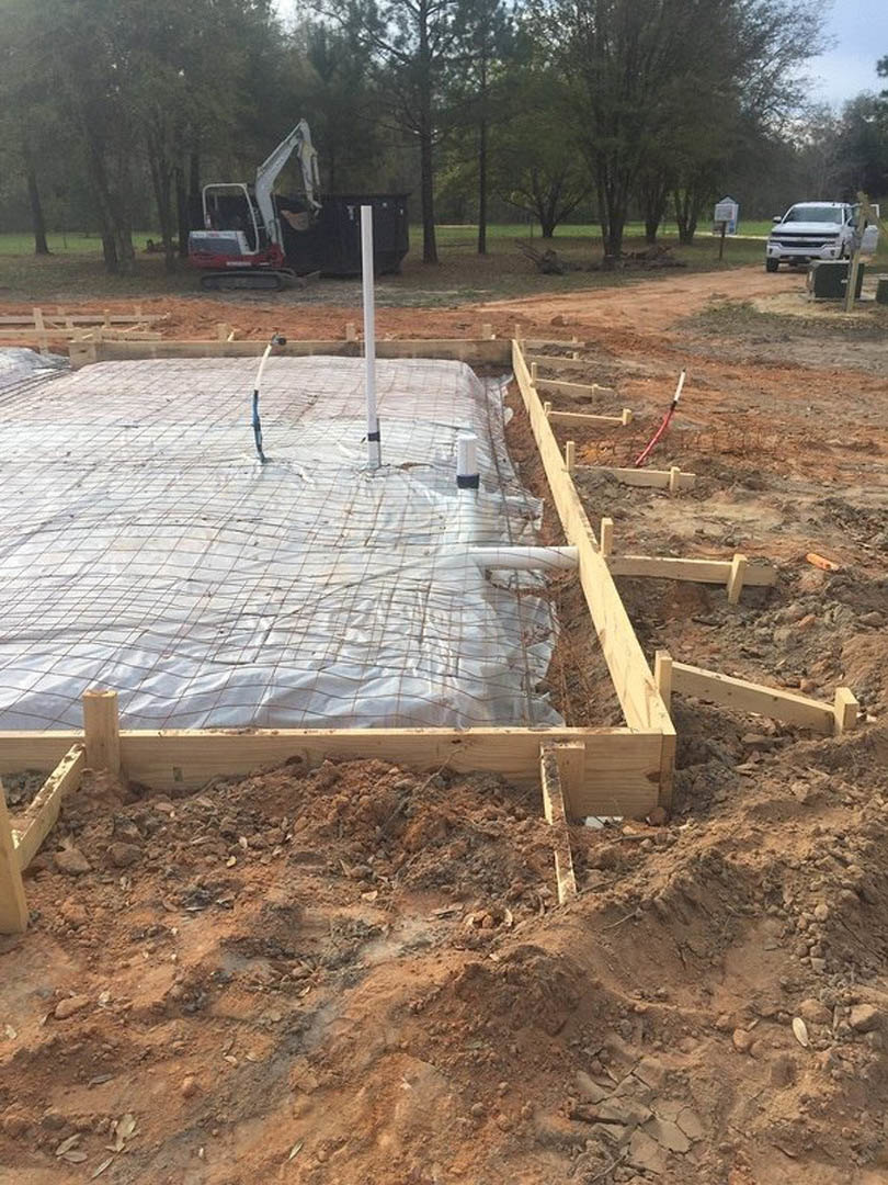 Foundation covered with white plastic sheet, wooden beams, and white pole with sign at residential construction site; white truck parked nearby, soil and trees in background.
