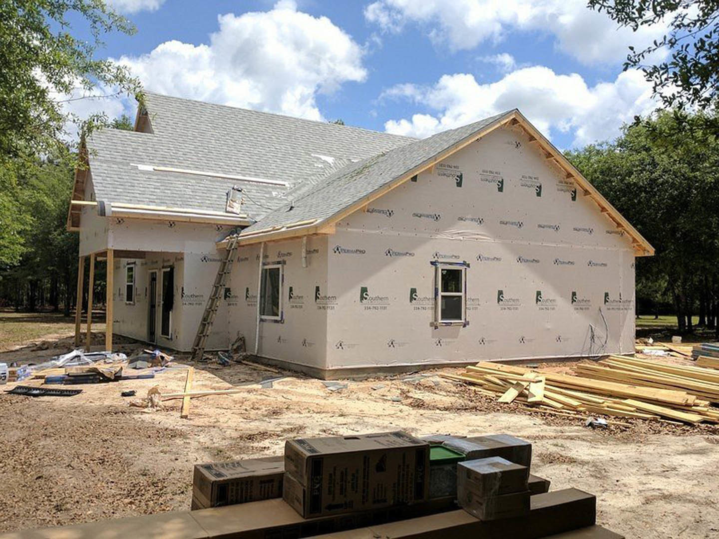 Partially built house with exposed wooden framing, white window frame, ladder leaning against wall, pile of lumber and stacked cardboard boxes on dirt ground, blue sky with
