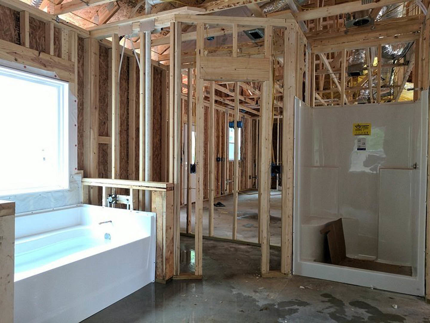 Bathroom under construction featuring exposed wooden framing, white bathtub, white shower enclosure with yellow caution sign, unfinished walls, and window with clear plastic