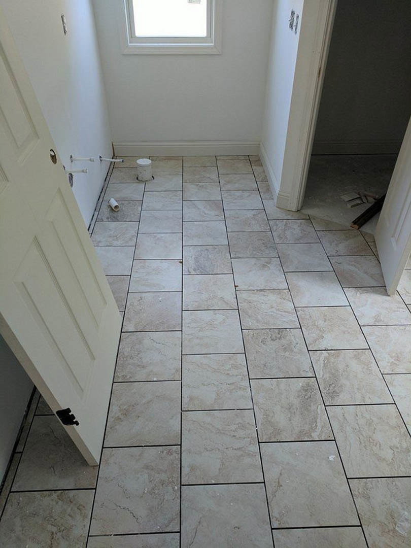 Tile flooring in a room with white plaster walls, white door, and a window; roll of toilet paper visible near the door, close-up details of grey tile surface.