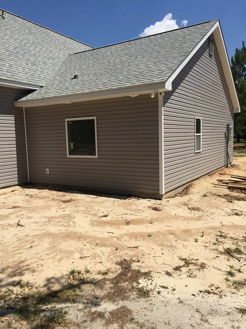 Modern home under construction with open door and window, dirt yard with scattered wood, patchy grass, and blue sky with white clouds