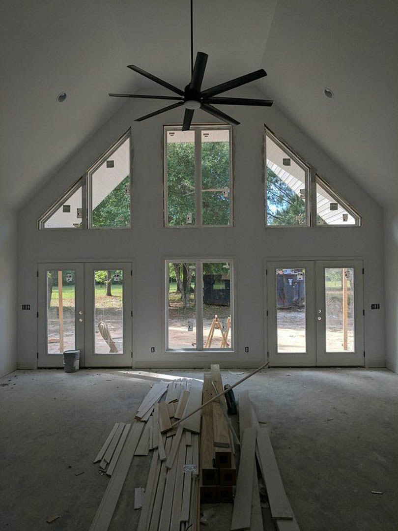 Spacious room featuring a white ceiling with ceiling fan, multiple windows and glass double doors, wood planks stacked on the floor, and natural daylight illuminating the interior.