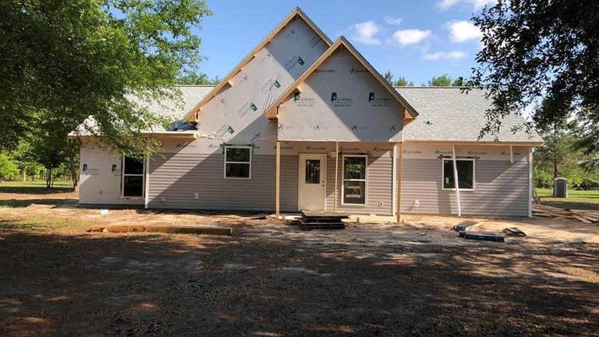 Partially built house surrounded by mature trees, exposed wooden framing and roof, dirt yard with wooden pallet, white framed windows and door visible on exterior