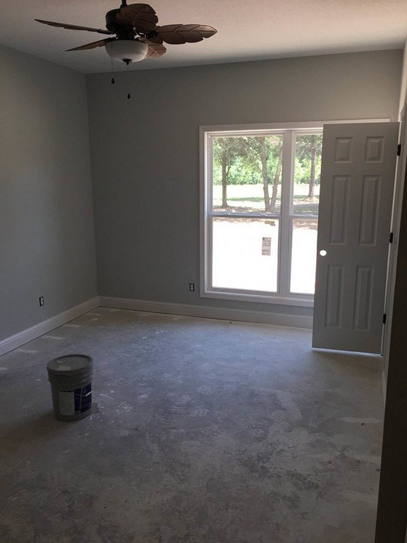 Unfinished room with concrete floor, white door featuring a hole, ceiling fan with light fixture, bucket of paint on the floor, and window showing trees outside