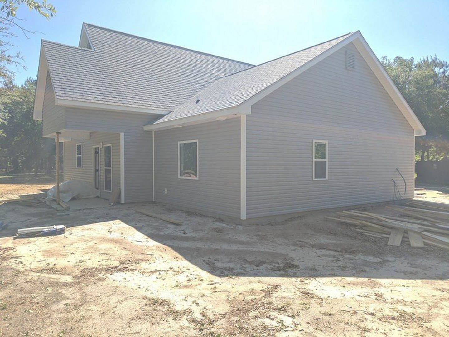 Partially built house with exposed framing and white window trim, sunlit exterior siding, dirt patch in front, blurred car on nearby road, trees and blue sky in background