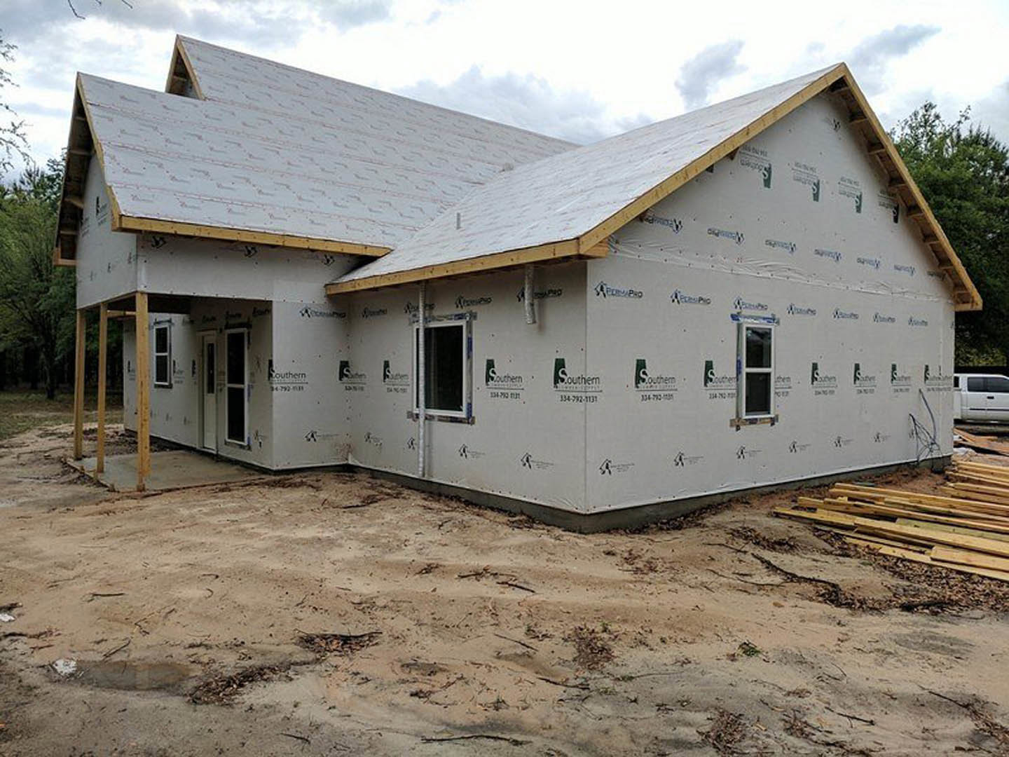 Framed custom home under construction with exposed wood beams, partially installed insulation, pile of lumber on dirt ground, white pickup truck parked roadside, cloudy sky