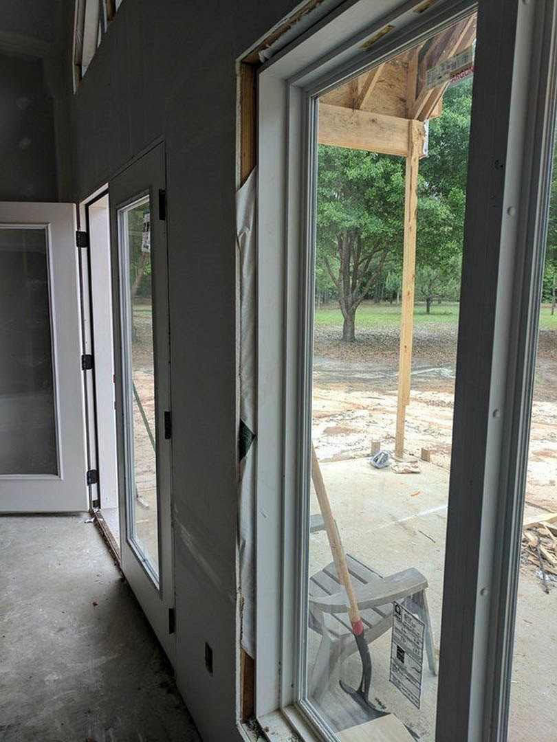 Modern living space featuring floor-to-ceiling glass doors, light wood flooring, and a single upholstered chair positioned near the window with views of a leafy tree outside.