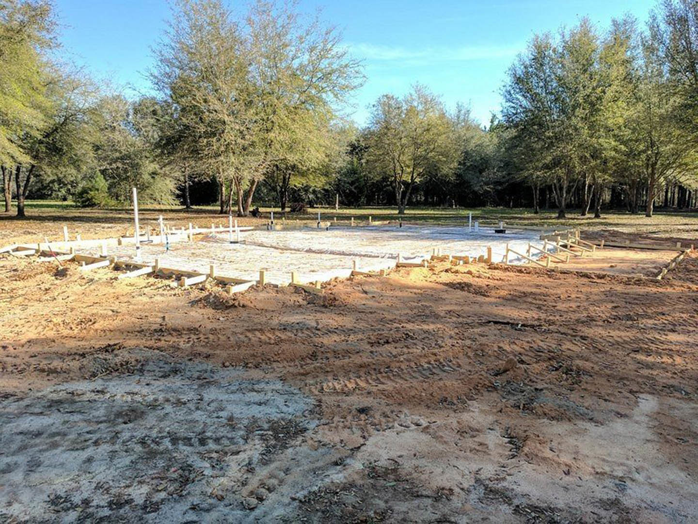 Dirt construction site with tire tracks, piles of soil, and snow patches; trees and wooden fence in background