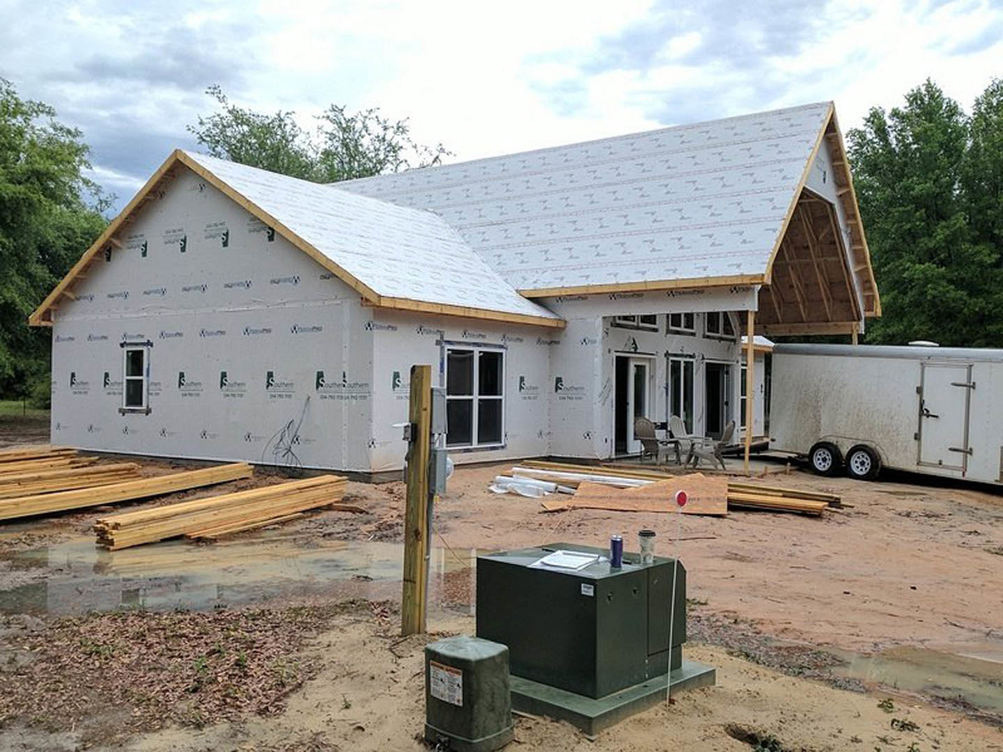 Partially built house with exposed framing and roof, construction trailer parked on dirt ground, scattered tools and materials, trees and cloudy sky in background