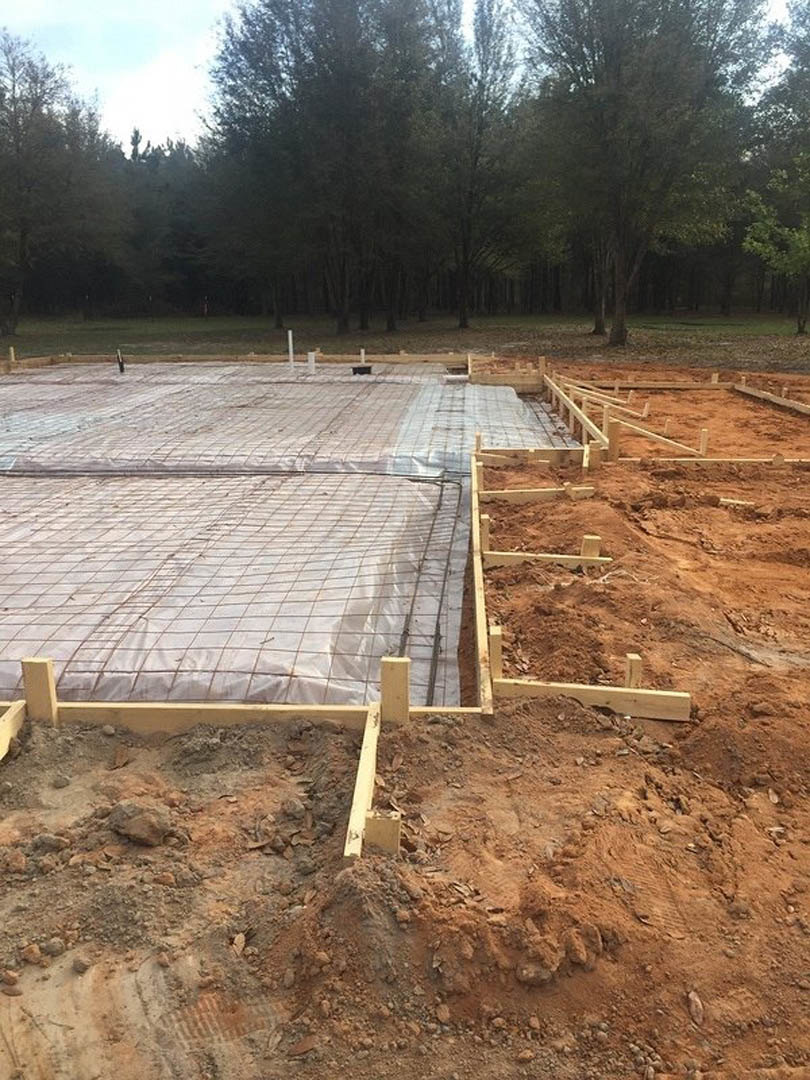Wooden beams and wire mesh forming a foundation on a dirt construction site, white tarp covering materials nearby, surrounded by forest trees.