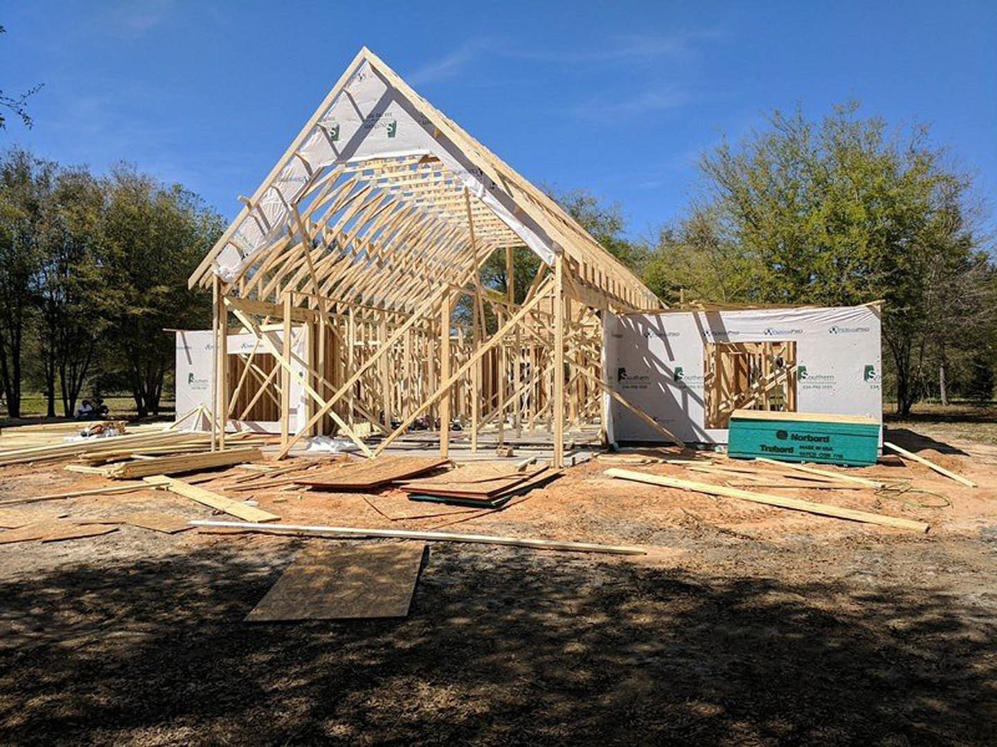 Wood-framed house under construction with exposed beams, unfinished walls, and dirt ground, surrounded by trees and cloudy sky