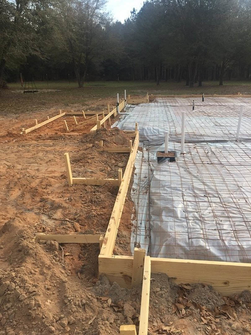 Framed foundation with exposed wood beams and metal bars, white tarp covering part of the site, soil and trees in background