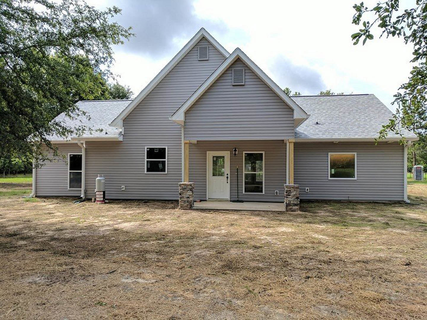 Two-story house with white siding, black-framed windows, and a white front door, surrounded by a green lawn and mature trees under a partly cloudy sky