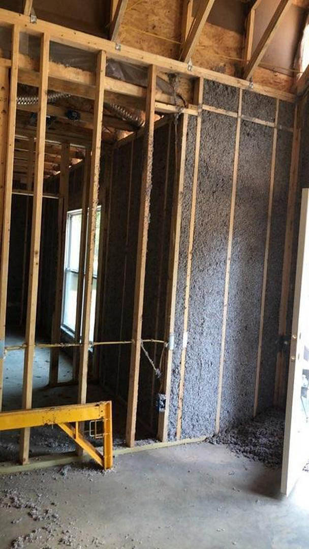 Yellow metal beam supported by wooden legs on concrete floor, unfinished interior with exposed lumber framing, window installed in wall, metal clip attached to wooden frame.