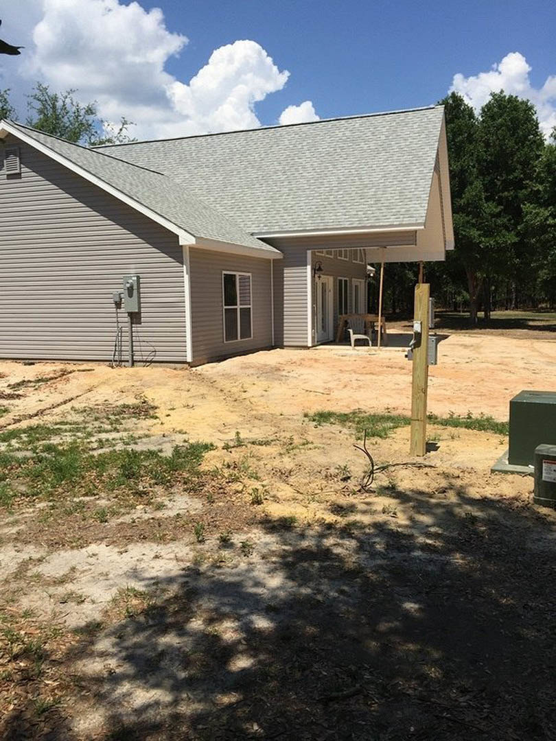 Two-story home with white-framed windows, attached garage, expansive paved driveway, leafy tree casting shadow on dirt ground, partly cloudy sky overhead