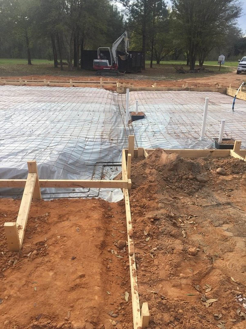 Wood-framed foundation on a dirt construction site surrounded by trees, white tarp covering part of the area, backhoe parked with open door, scattered leaves on soil.