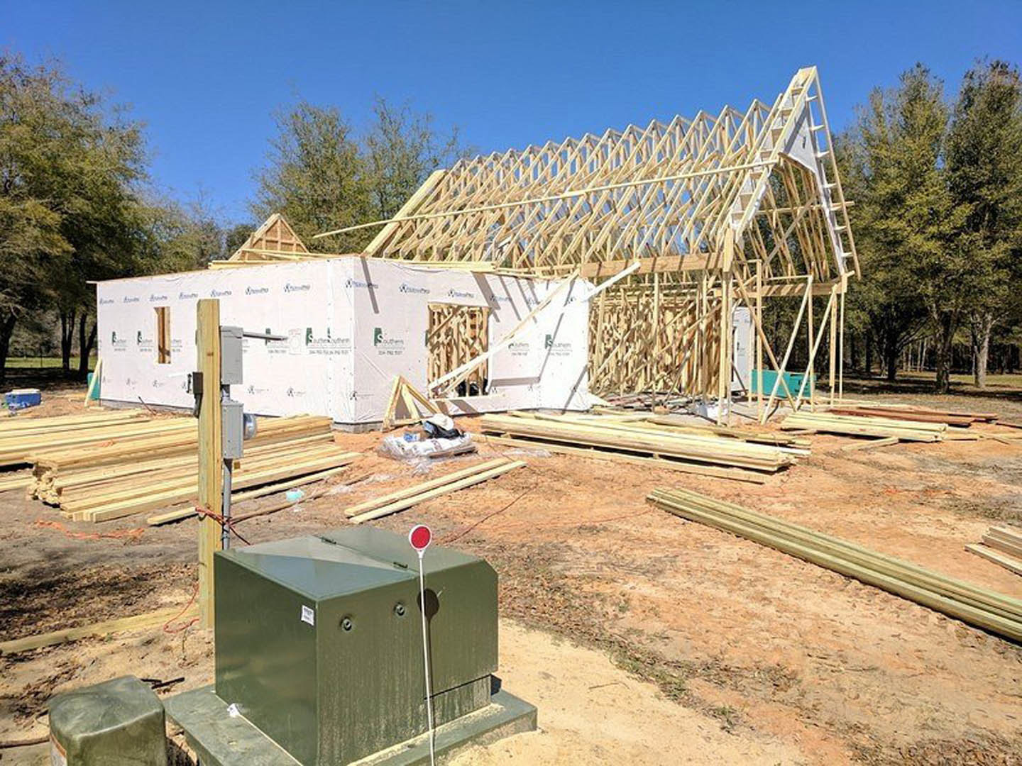 White square house under construction with exposed wood framing, partially finished roof, green utility box with sign in foreground, metal pipe, and leafy tree nearby.